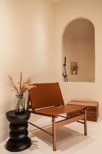 A minimalist indoor lounge area featuring a leather and wooden chair, a vase with dried plants on a black modern table, and a