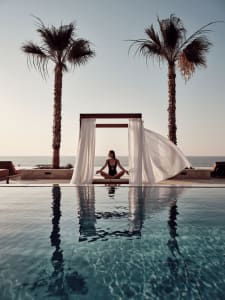 A woman in a minimalist setting sitting on a sun drape by an infinity pool with tropical surroundings