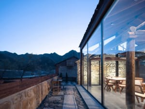 Outdoor seating area with stone walls, glass walls, and concrete patio at dusk, overlooking mountains.