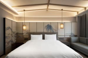 A modern minimalist bedroom featuring a large bed with white bedding, a grey sofa, and a wall with traditional Chinese artwor