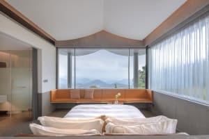 A modern bedroom with a white bed, orange seating area, and large windows overlooking mountains.