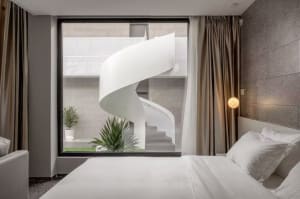 A minimalist bedroom with concrete walls and a white bed, large window overlooking a spiral staircase and greenery.