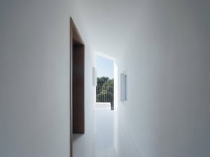 Minimalist interior design with white walls, ceiling, and floor featuring a corridor with a door and a window showing a balco