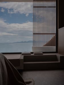 Minimalist resort room featuring stone cladding and wood-paneled walls overlooking the ocean, with a bathtub and books.