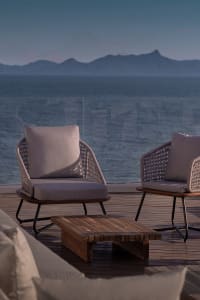 Outdoor seating arrangement featuring wooden furniture and cushions near the ocean with mountains in the background.
