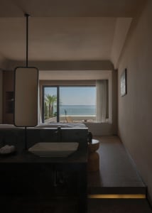 Modern bedroom with concrete walls and floor, large window overlooking the ocean, and minimalist furniture.