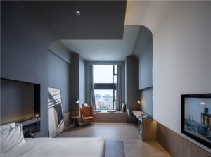 A modern minimalist bedroom featuring a large bed with a white comforter, a white accent chair, and a large window with citys