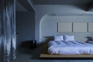 Minimalist bedroom with white bedding on a concrete platform bed, three white square paintings on the wall, and concrete floo