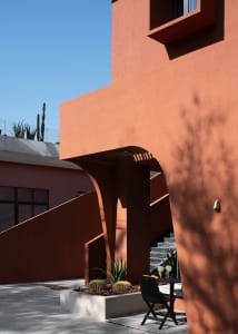 Modern exterior of a clay building with cactus and a black table and chair.