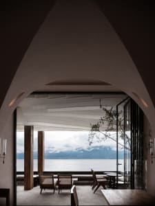 Modern restaurant interior with wooden furniture, marble floor, and scenic view of mountains and water.