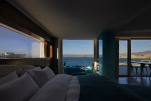 A hotel room interior featuring a glass wall with a view of the ocean and cityscape.