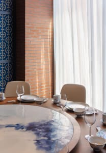 A round wooden table set in an industrial restaurant interior with white curtains and a brick wall.