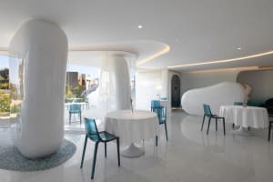 A minimalist restaurant interior featuring white walls, glass tables, and concrete columns.