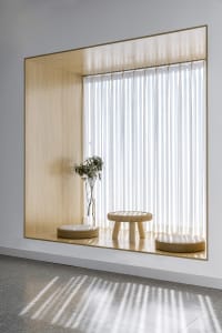 A minimalist window seat with white walls, featuring a vertical blind curtain, two cushioned stools, and a small plant in a v