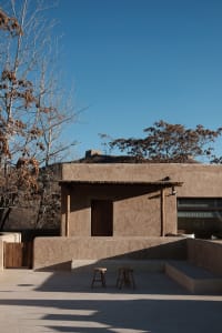 A minimalist clay exterior building with a wooden door and two stools in front of it.