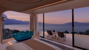 Modern hotel bedroom featuring a glass bathtub overlooking a scenic ocean view at sunset.