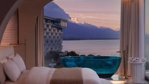 Modern hotel suite featuring a glass bathtub overlooking a lake at sunset.
