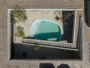 A modern pool with a circular design, surrounded by a stone deck and concrete elements, featuring trees and a metal railing.