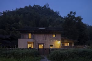 A house with a concrete exterior illuminated at night, surrounded by dark green foliage and tall trees in the background.