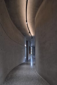 Modern concrete corridor interior with curved walls and floor, minimalistic lighting, and a view into another room.
