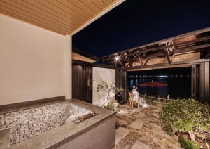 A luxurious residential patio at night, featuring an outdoor hot tub with a metal-tile surround, a concrete deck, and a sceni