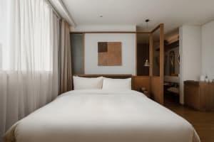 A modern minimalist bedroom with a large bed featuring white fabric, surrounded by wooden elements and a large window with sh