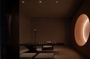 A minimalist spa lounge featuring dark interior with soft textiles, wooden walls, and stone floor.