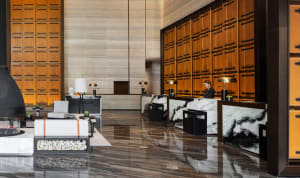 A modern hotel lobby featuring marble and concrete finishes, with a receptionist desk and seating area.