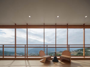 Two wicker chairs and a black table with a glass top overlooking a scenic landscape through large glass windows.