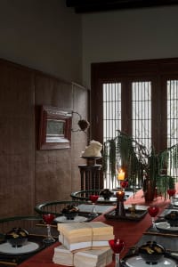 A beautifully arranged dining table with a red tablecloth, red wine glasses, black and gold bowls, and stacks of books. The r