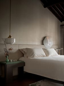 A minimalist bedroom featuring a white bed with a simple headboard, a side table with a vase and tea set, and a pendant light