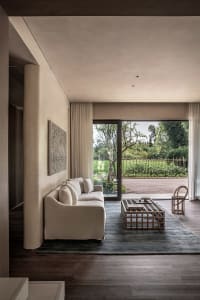 A modern living room with a beige sofa, a wooden coffee table, and a large window overlooking a green outdoor area.