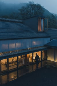 A modern house with a glass exterior at night with lights on and a person walking outside.