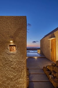 A minimalist building exterior with concrete walls and a wooden door, illuminated at dusk with a scenic ocean view.