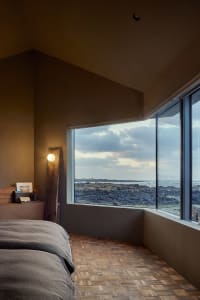 A modern bedroom featuring a wooden floor, a bed with a gray blanket, and a window overlooking a rocky coastline.