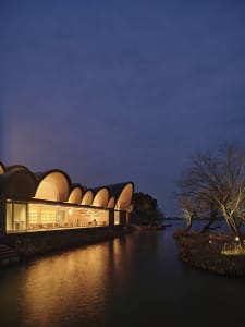 Modern residential house with glass facade overlooking a lake during dusk.