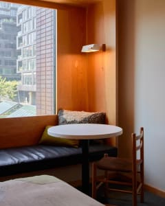 A minimalist window seat with a concrete table and wooden chair, featuring warm lighting and a view of surrounding buildings.