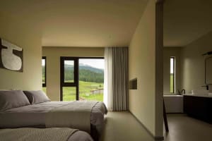 A modern minimalist bedroom featuring neutral-toned decor, a cozy bed, and a floor-to-ceiling window overlooking a green land