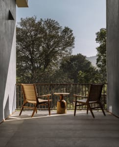 Two wooden armchairs and a wooden side table on a balcony overlooking forested mountains.