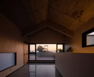 Modern interior with concrete walls and floor, large windows showing a sunset view.
