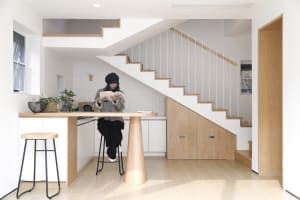 A modern kitchen with white oak wood cabinets, a white countertop, and a white staircase in the background. A woman is sittin