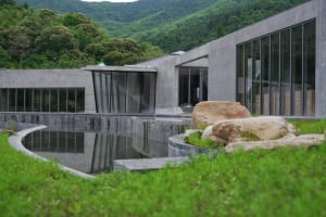 A modern building made of concrete with a pool in the foreground, surrounded by greenery and large rocks.