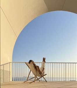 A person relaxing on a deck chair with their feet up on a balcony overlooking the ocean.