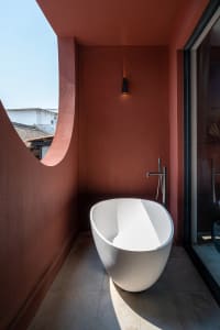 Minimalist white ceramic bathtub in a red-walled bathroom with a circular cut-out in the wall and open glass door.