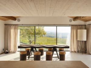 Modern dining room with minimalist design featuring a wooden table and stools, large window overlooking a scenic vineyard.