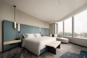 A modern hotel room featuring a bed with a white comforter and a blue headboard, overlooking a cityscape through a large wind