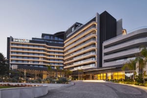 Paved driveway leading to the entrance of a modern hotel building with illuminated balconies and landscaping.