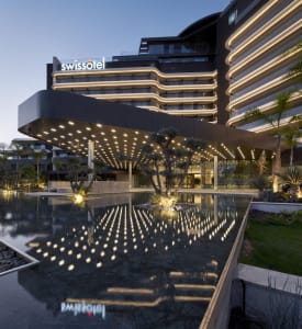 Modern hotel building with metallic facade and glass windows, illuminated at dusk with reflected lights in a water feature.