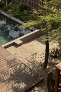 Swimming pool deck surrounded by greenery with concrete, sand, and wooden elements.