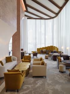 A modern hotel lobby featuring plush, cushioned seating areas with large windows and a high, curved ceiling.